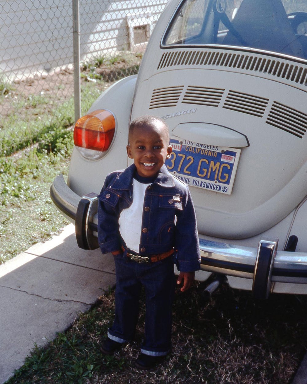 Color photo of a young boy standing in front of a white Volkswagen Beetle in Los Angeles, 1977. He wears a matching dark denim jacket and jeans with a white turtleneck and a brown belt with a large brass buckle, and grins broadly at the camera. A California license plate reading "372 GMG" is visible behind him.