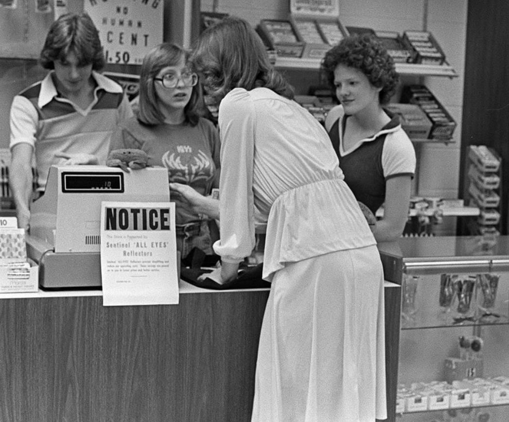 Black-and-white photo of a customer leaning over a retail counter toward two young female cashiers at a store in the Poconos, Pennsylvania, in 1977. The cashier on the left wears glasses and a KISS t-shirt; the cashier on the right looks on with a slight smile. A "Notice" sign and a cash register sit on the counter between them. Candy and merchandise are stacked on shelves behind the counter.