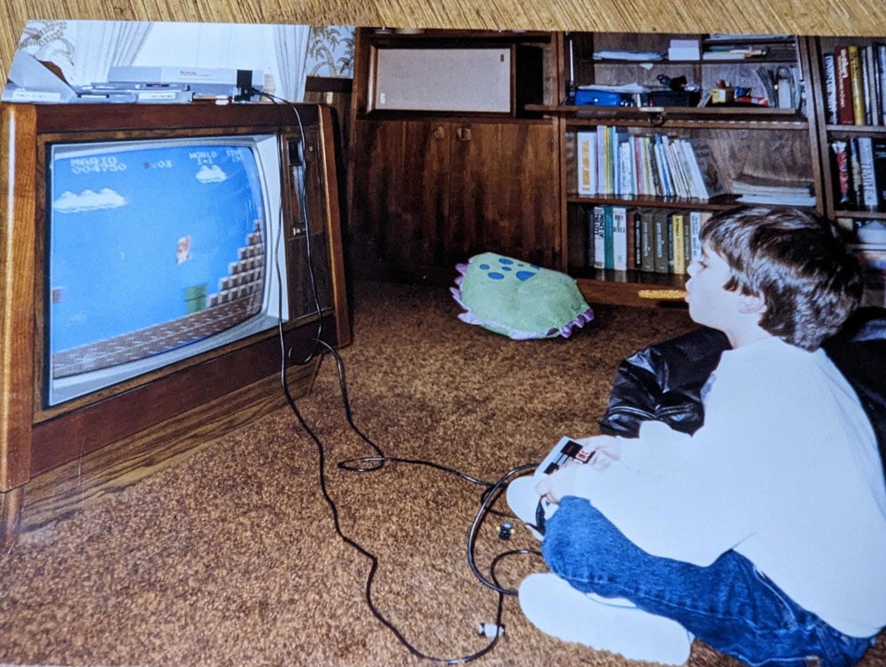 A young boy sits cross-legged on shag carpet playing Super Mario Bros. on a Nintendo Entertainment System connected to a large wood-paneled television, with a stuffed animal and bookshelf visible behind him.