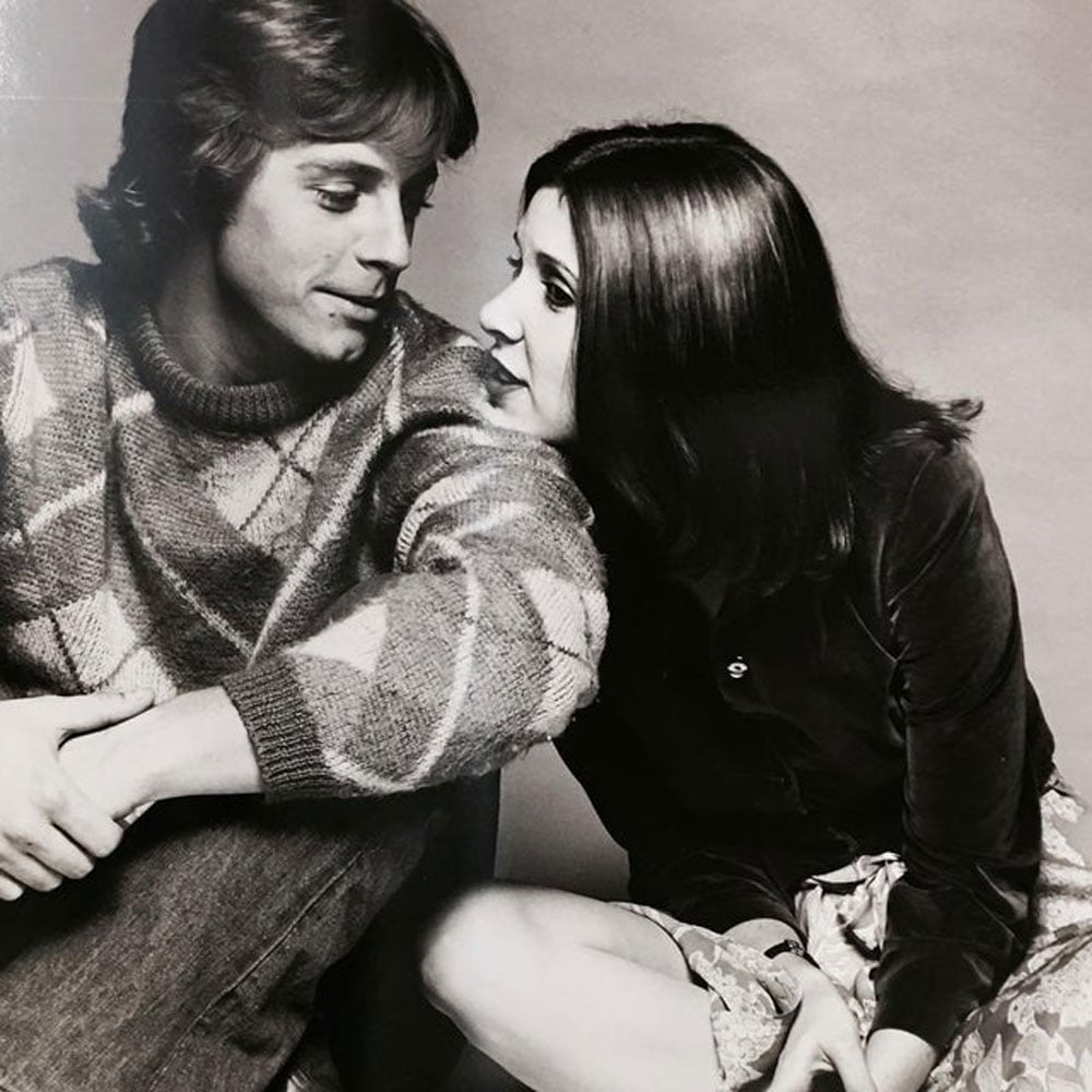 Black-and-white photo of Mark Hamill and Carrie Fisher seated close together during a 1977 Star Wars promotional shoot. Hamill wears a patterned sweater and looks at Fisher; she leans toward him, her long dark hair falling forward. The background is a plain studio backdrop.