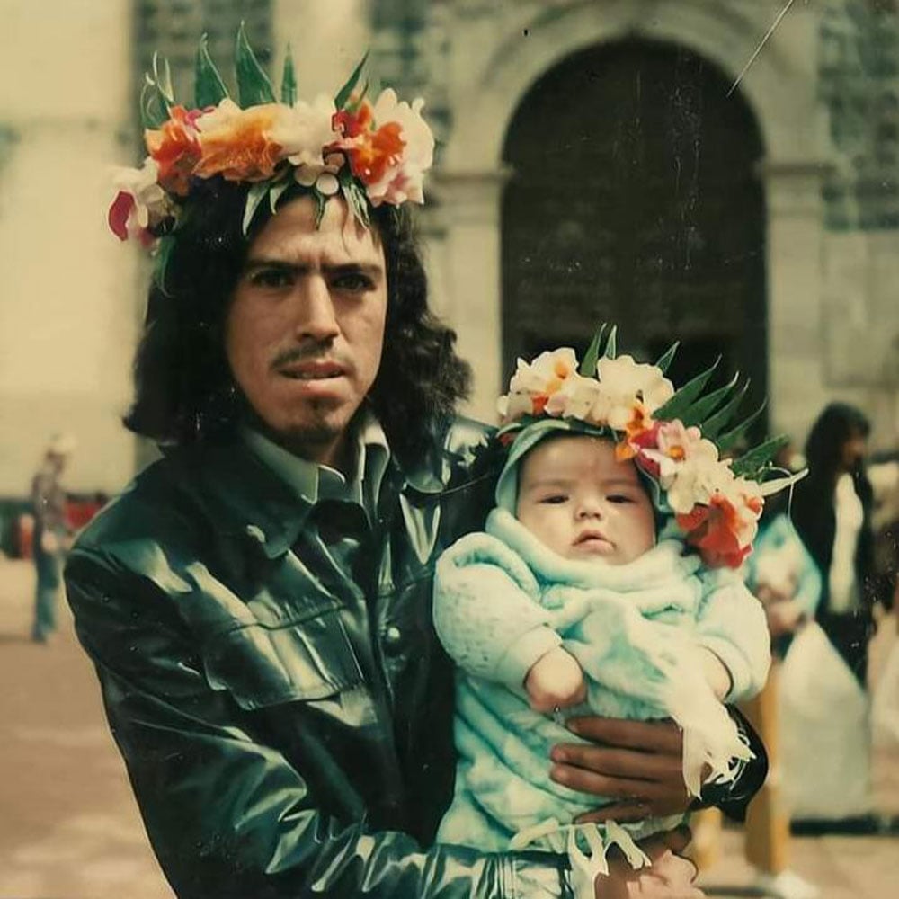 Color photo of a young father holding a newborn baby in Mexico City in 1977. Both wear matching flower crowns made of orange, pink, and white blossoms and green leaves. The father wears a dark leather jacket; the baby is bundled in a light blue blanket. A stone cathedral arch is visible behind them.