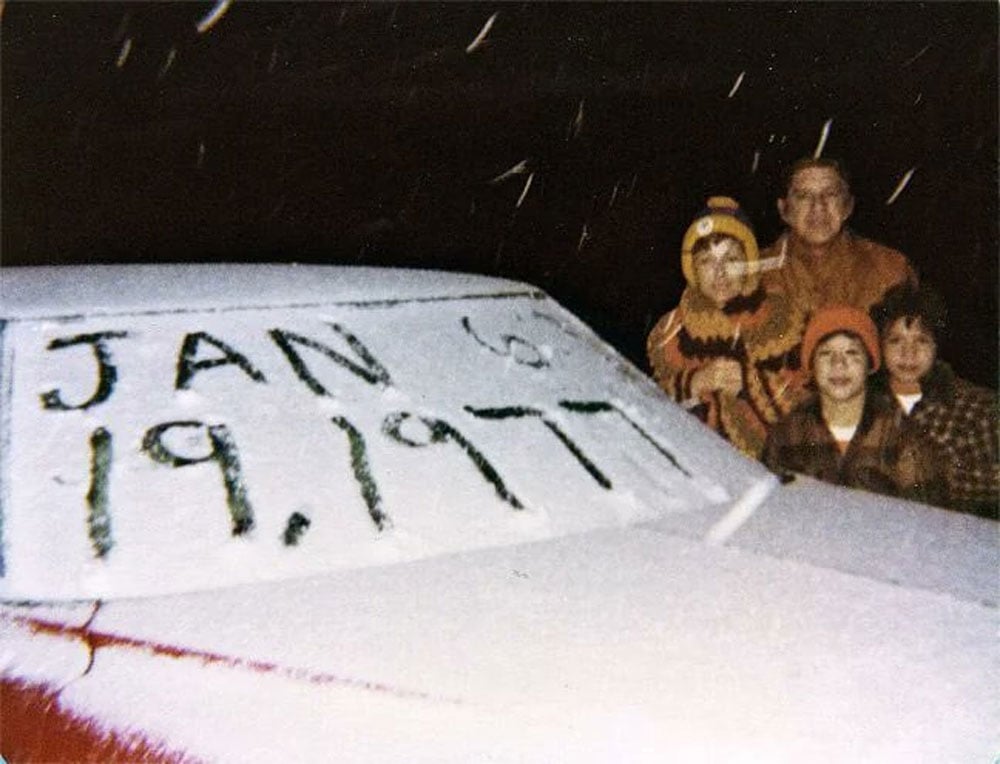 Color photo of a family posing behind a snow-covered car hood at night in Miami, Florida. The date "Jan 19, 1977" is written in the snow on the windshield. An adult and three children stand bundled up behind the car, grinning, as snow falls around them.