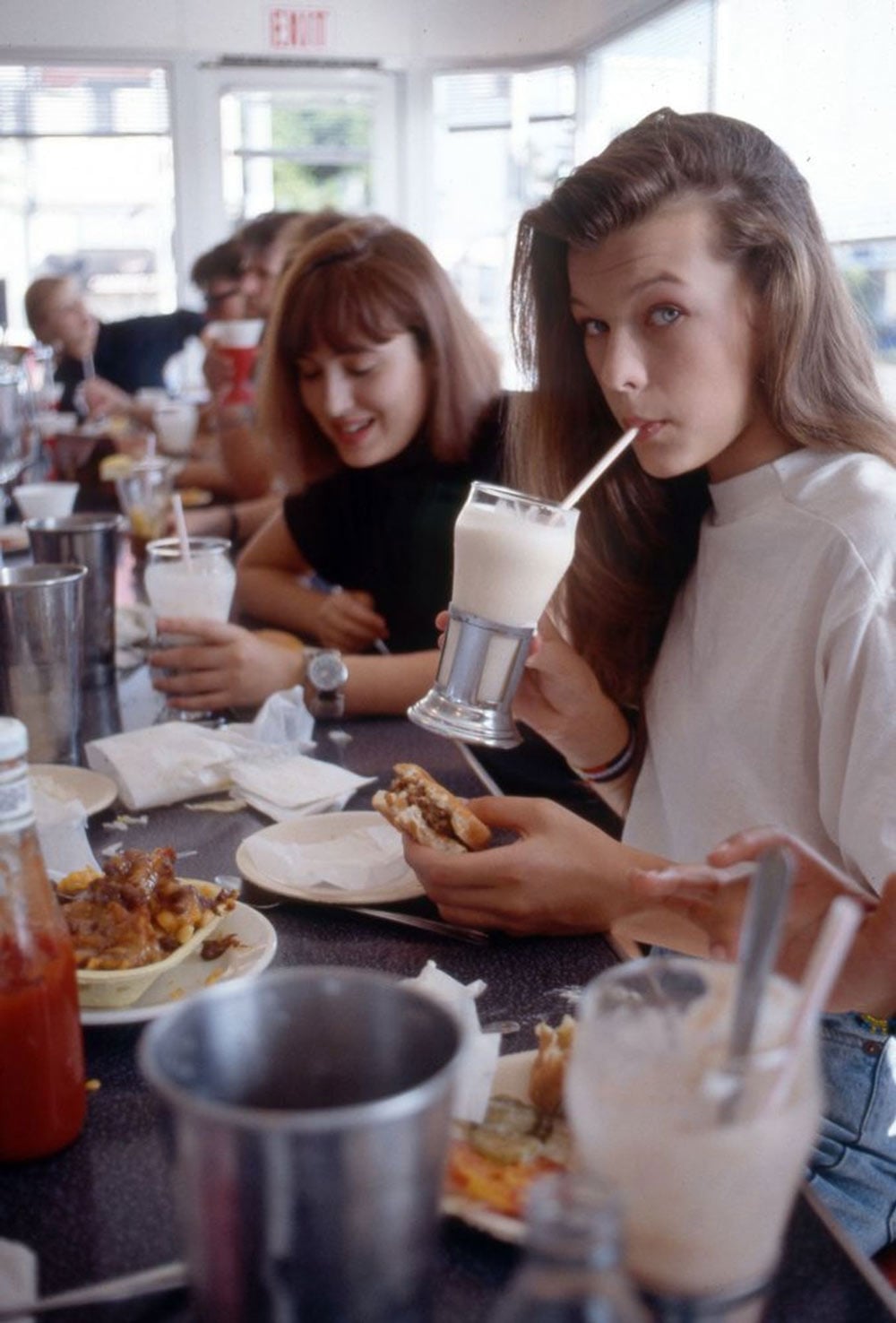 A young teenage girl sips a milkshake at a diner counter while holding a burger, with other diners visible along the counter behind her.