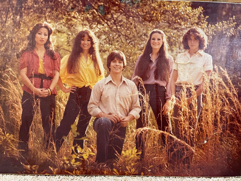 Color photo of five young siblings posing together in a field of tall golden grass in 1977. Four women and one young man stand together among autumn foliage. They wear earth-toned blouses and dark pants; several have long dark hair.