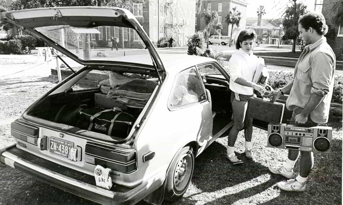 Black and white photo of a woman unpacking her car and handing a case of cassettes to a man holding a boom box