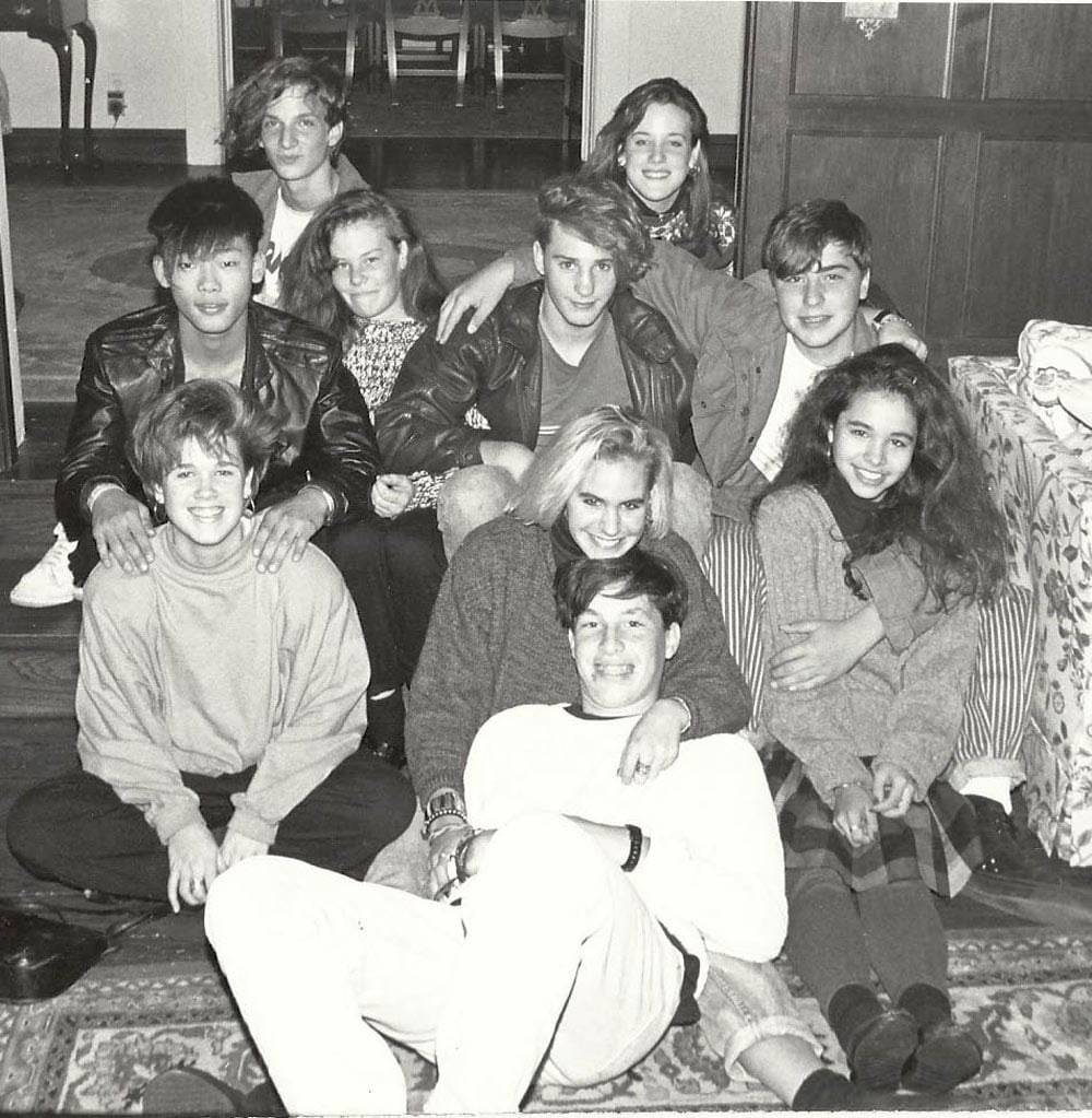 A group of about ten teenagers crowd together on the floor and a couch in a living room, smiling at the camera in a black and white photo.