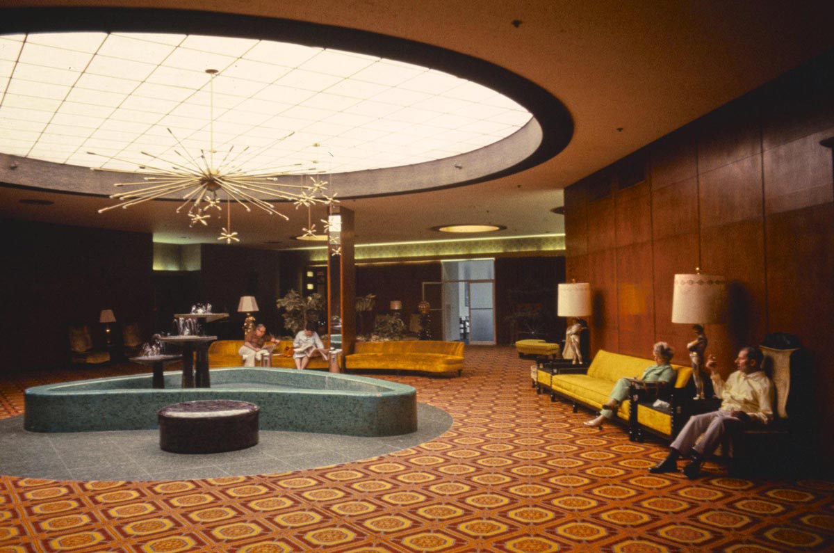 Color photo of the ornate lobby of the Nevele Hotel in Ellenville, New York, in 1977. A large oval skylight illuminates the space from above, beneath which hang Sputnik-style chandeliers. A kidney-shaped turquoise fountain sits in the center of a boldly patterned red and gold carpet. Several guests sit on gold velvet sofas along the walls, and a small indoor fountain is visible in the background.