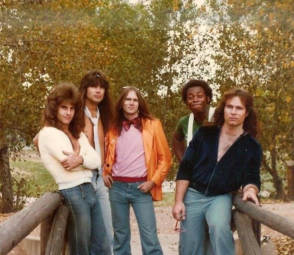 Color photo of five members of the band Orphan Annie posing on a bridge in Denver, Colorado, in 1977. All five wear jeans and casual tops; trees are visible in the background.