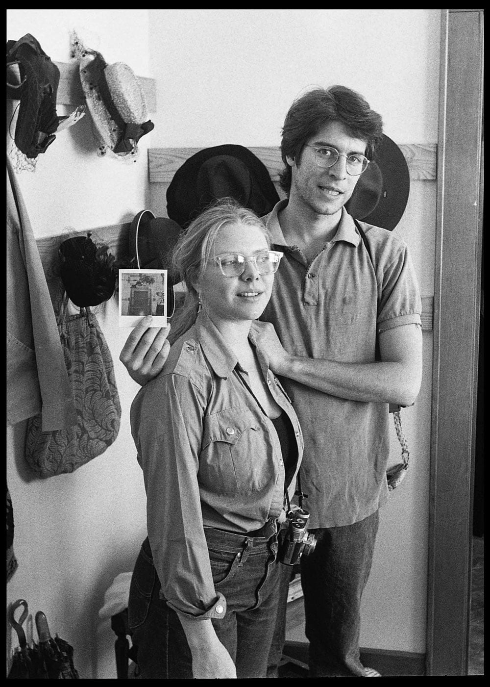 Black-and-white photo of a young couple standing together in a room with a hat rack on the wall behind them. The woman wears large clear-framed glasses and a button-up shirt, with a film camera hanging from her hip. The man behind her wears wire-rimmed glasses and a polo shirt, holding up a small Polaroid photo with one hand and resting the other on her shoulder.