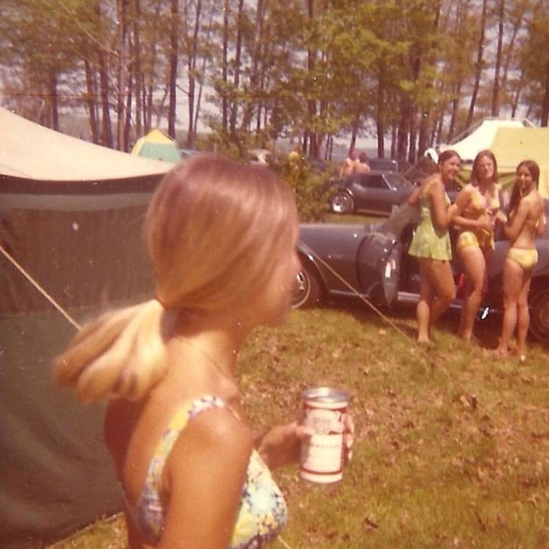 Color photo of a lakeside campsite party in 1977. Several young women in bikinis stand near parked cars and canvas tents on a grassy bank, with pine trees and a lake visible in the background. A woman in the foreground holds a can of beer, her hair caught mid-turn by the camera.