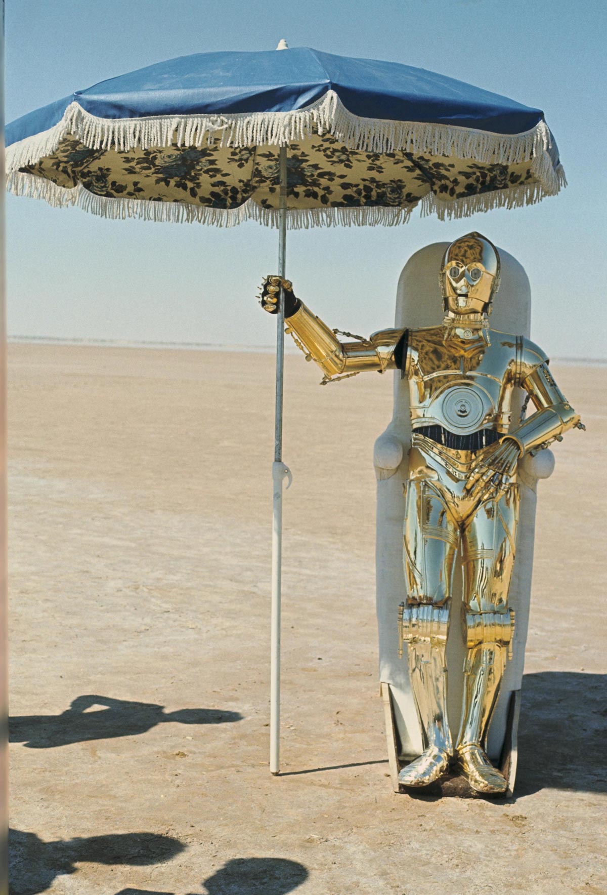 Color photo of actor Anthony Daniels in the C-3PO costume standing in the Tunisian desert holding a fringed blue beach umbrella, leaning against a white chair. The gold protocol droid costume gleams in the sun against a flat sandy landscape and pale blue sky.