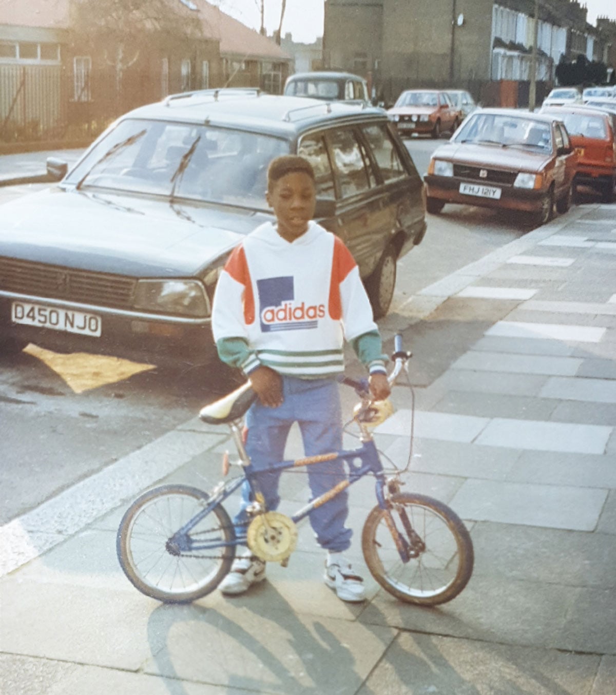 A young boy in an Adidas hoodie and jeans stands on a sidewalk next to a Raleigh Burner BMX bike, with parked cars and a row of brick buildings visible on a British street behind him.