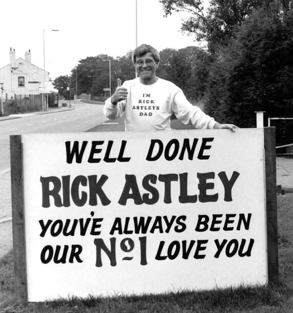 A man wearing a sweatshirt reading "I'm Rick Astley's Dad" stands behind a roadside sign that reads "Well Done Rick Astley, You've Always Been Our No. 1, Love You," giving a thumbs up.