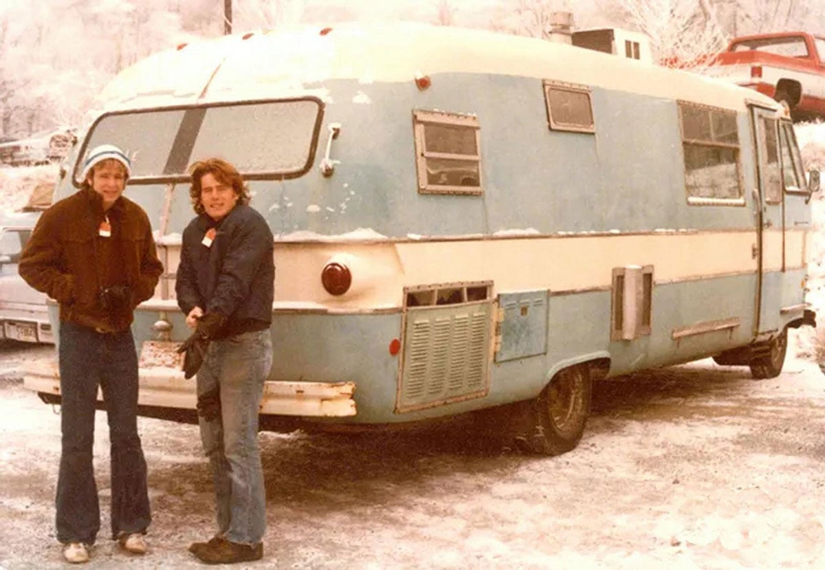 Color photo of two young people standing in front of a weathered blue and white RV in a snowy parking lot in 1977. Both wear winter jackets; the person on the left has on a knit hat. Ice-covered trees and a red truck are visible in the background.