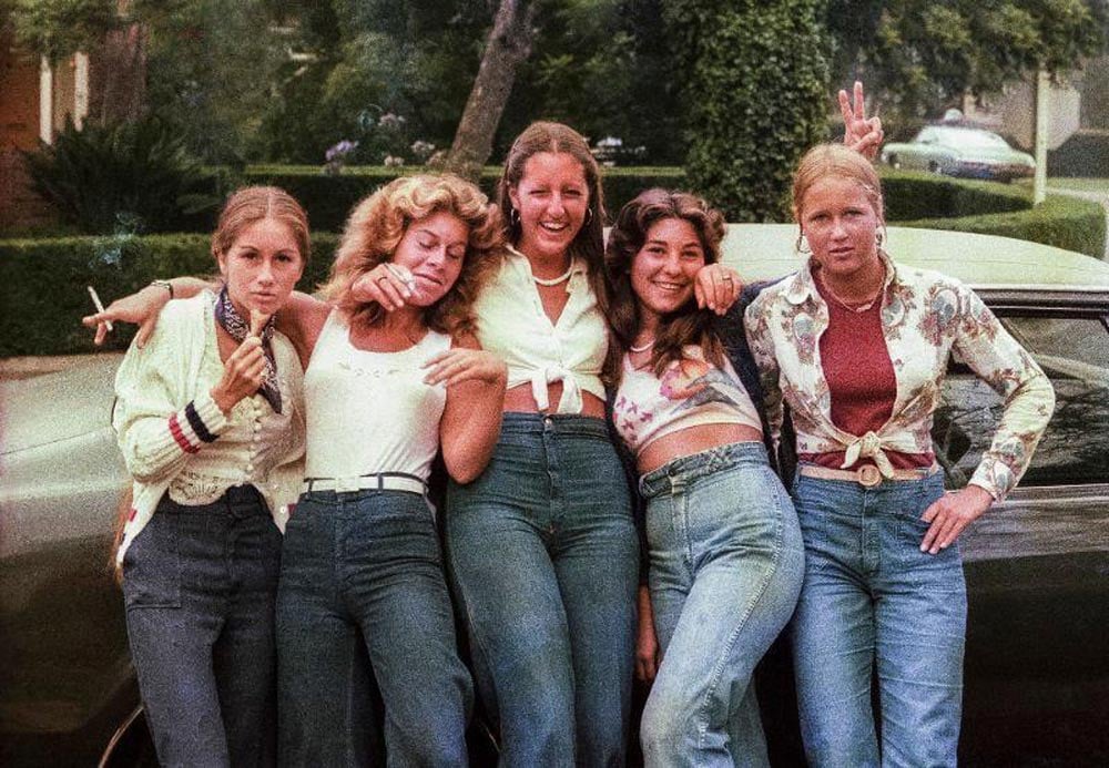 Color photo of five teenage girls leaning against a car on a tree-lined suburban street in San Diego in 1977. They wear high-waisted jeans and various casual tops including a tied blouse, a floral shirt, and a cardigan. One makes a peace sign and another rests her head on a friend's shoulder. Green hedges and parked cars are visible behind them.