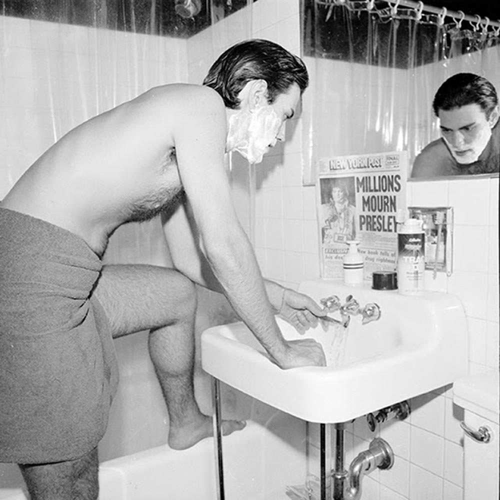 Black-and-white photo of a shirtless man leaning over a bathroom sink to shave, his face covered in shaving cream. Propped against the mirror is a copy of the New York Post with the front page headline reading "Millions Mourn Presley." Toiletries line the sink beside it.