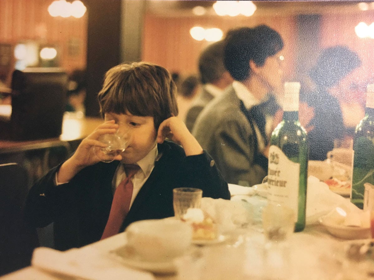 A young boy in a suit and tie sips from a wine glass at a restaurant table, with a wine bottle and other glasses visible nearby and adults seated behind him.