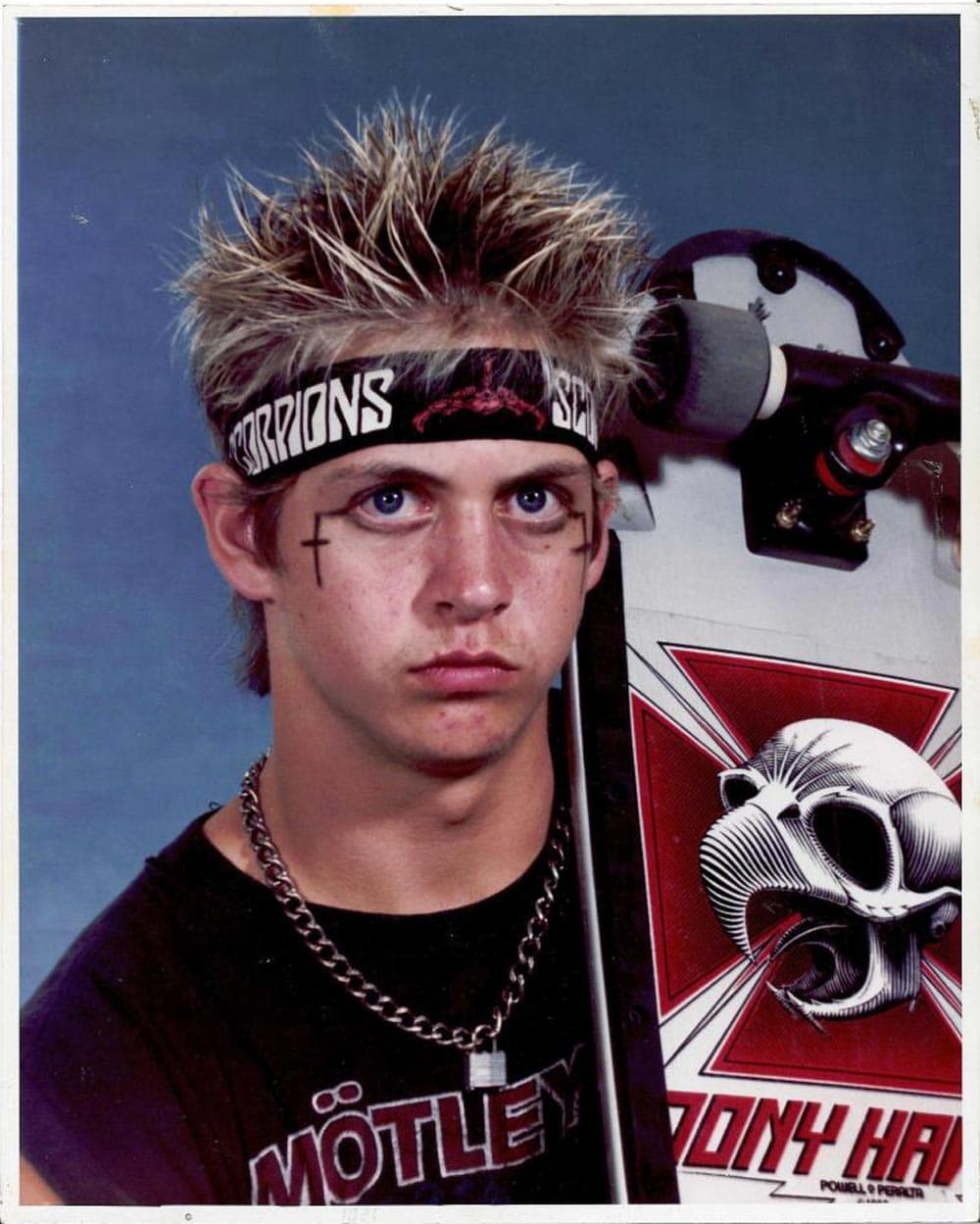 A kid with spiky hair and cross painted on his cheeks holding his skateboard for a school photo