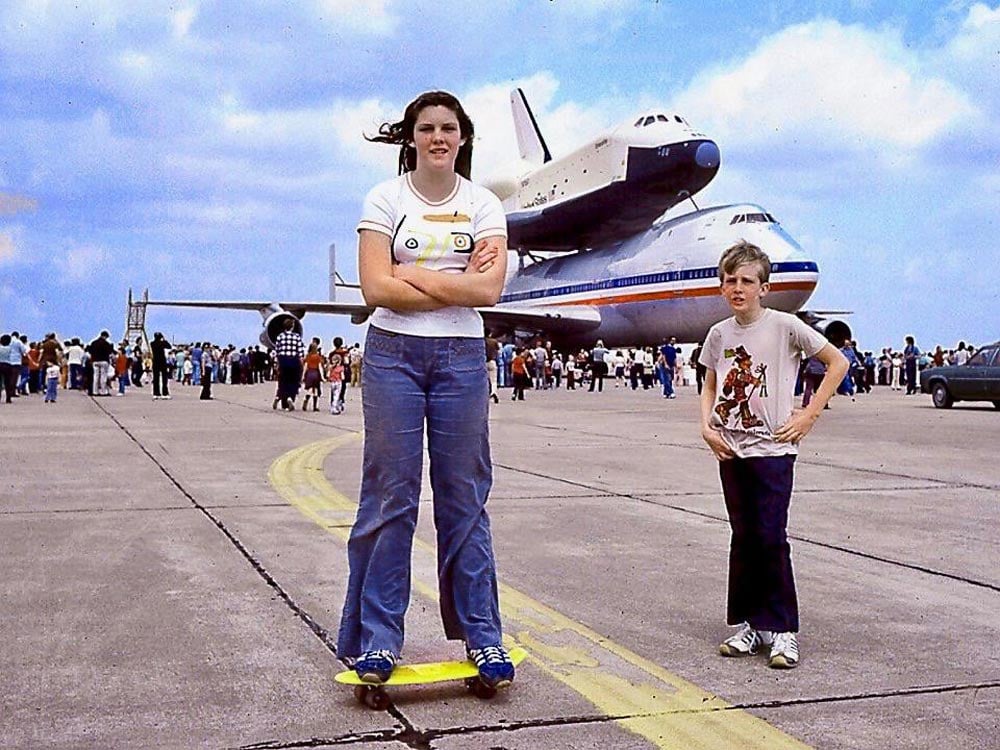 Color photo of a teenage girl standing on a yellow skateboard and a younger boy posing together on an airport tarmac in 1977, with the Space Shuttle Enterprise mounted atop a Boeing 747 visible behind them. A large crowd of spectators lines the tarmac in the background beneath a bright blue sky.