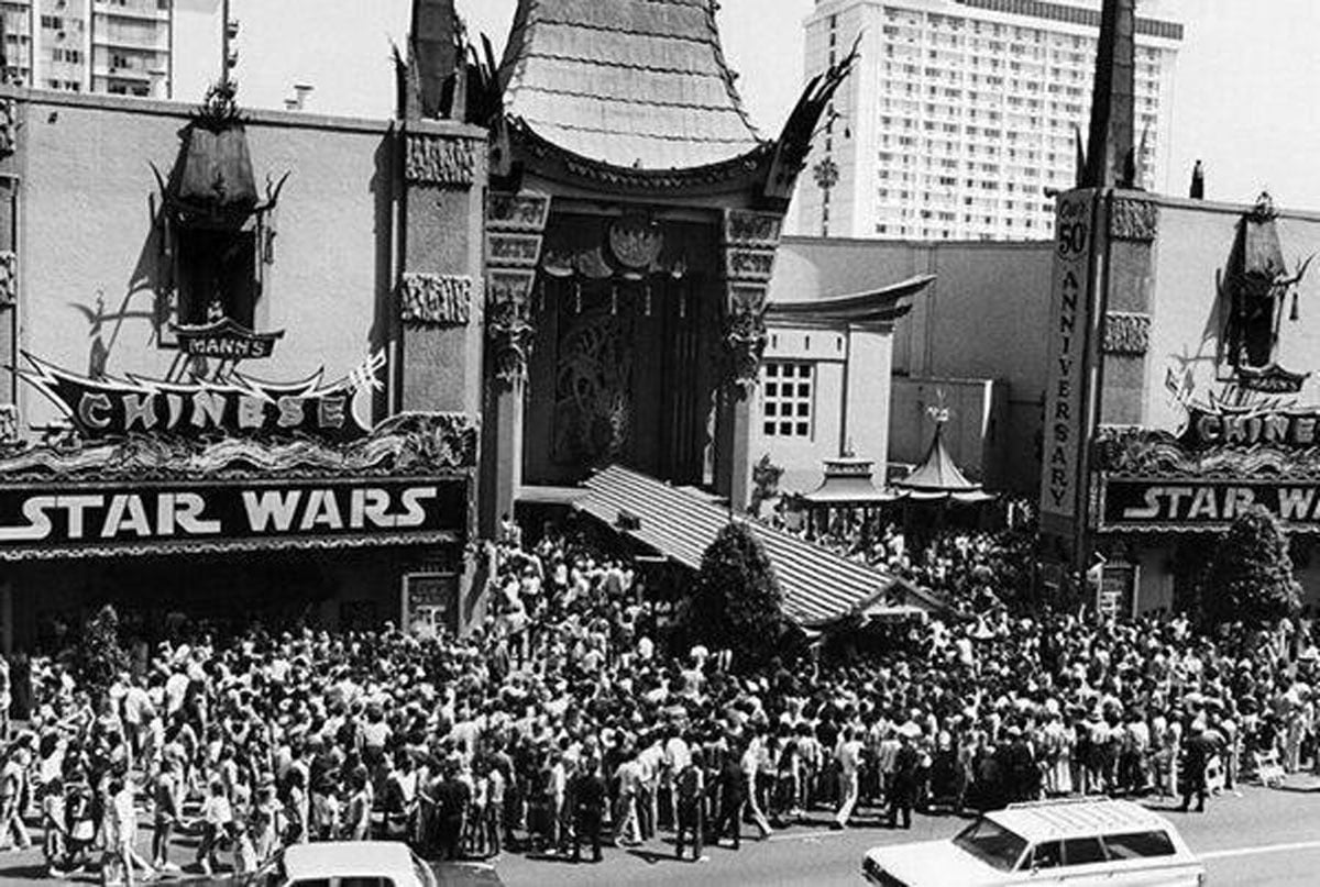 Black-and-white aerial photo of a massive crowd filling the sidewalk and street outside Mann's Chinese Theatre in Hollywood on opening day of Star Wars, May 25, 1977. The theater's marquee reads "Star Wars" on both sides, and a "50th Anniversary" sign is visible on the right. The crowd extends as far as the frame allows.