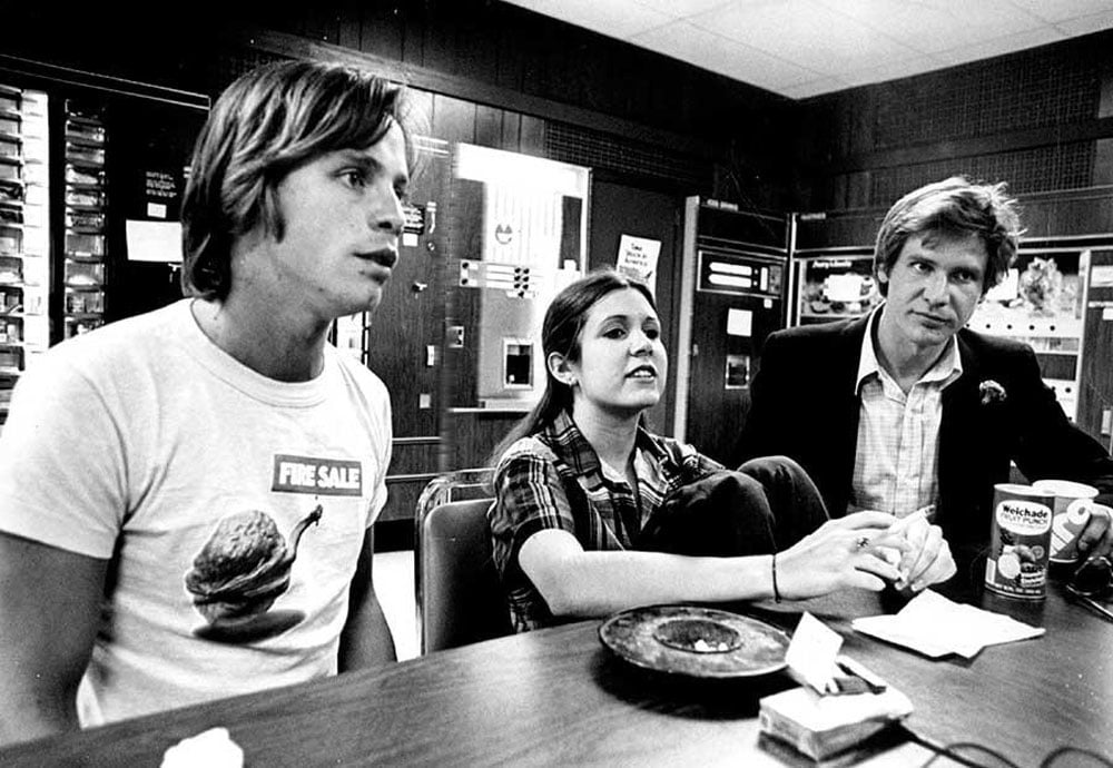 Black-and-white photo of Mark Hamill, Carrie Fisher, and Harrison Ford seated at a table during a press interview in Denver, 1977. Hamill wears a "Fire Sale" t-shirt and leans toward someone off-camera. Fisher sits in the middle, speaking. Ford sits on the right in a sport coat, holding a cup, with a can of Welchade Fruit Punch on the table in front of him. Vending machines line the wall behind them.