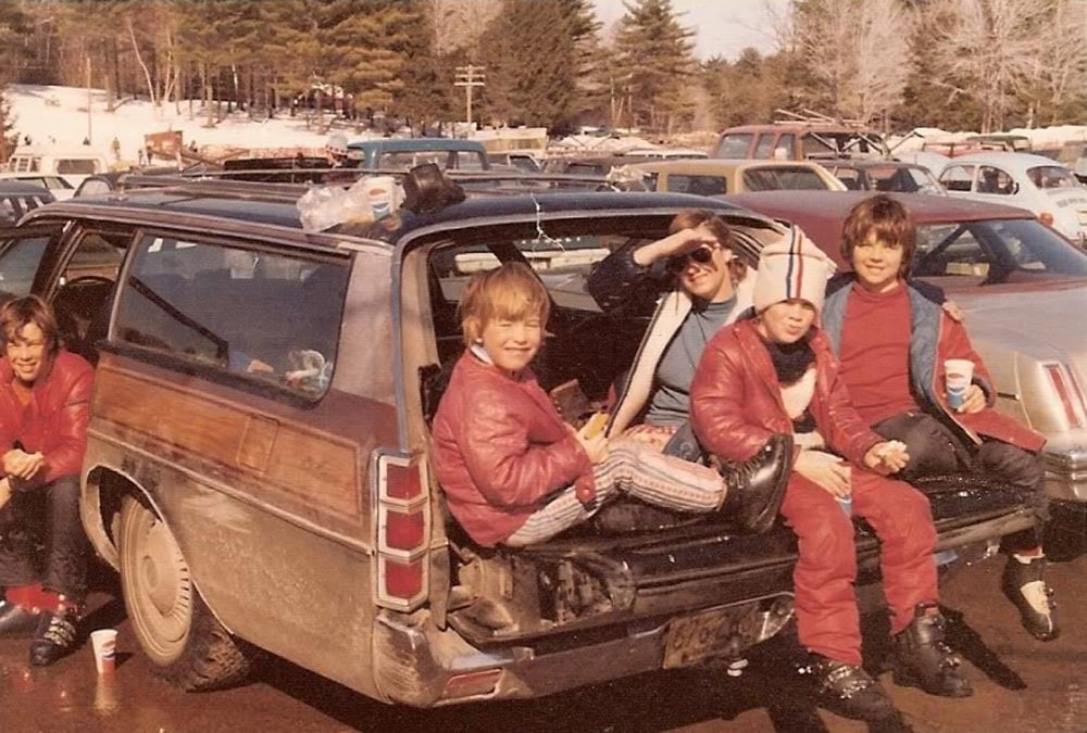 Color photo of four children and an adult sitting in the open back of a wood-paneled station wagon in a snowy ski mountain parking lot in 1977. The kids wear red ski jackets and ski boots and hold drinks and food. Pine trees and rows of parked cars are visible in the background.
