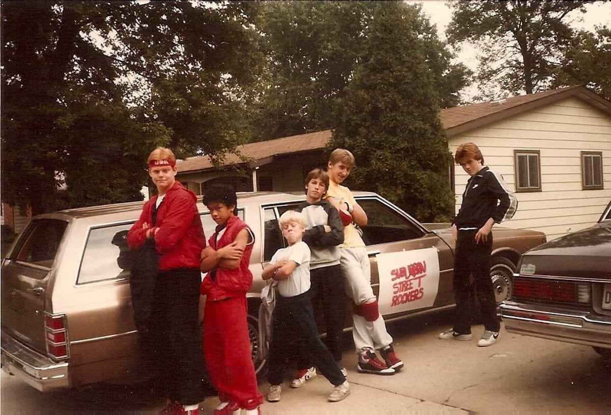 Six kids striking a pose in a driveway next to a station wagon with a sign tapped on it that reads "Suburban Street Rockers"