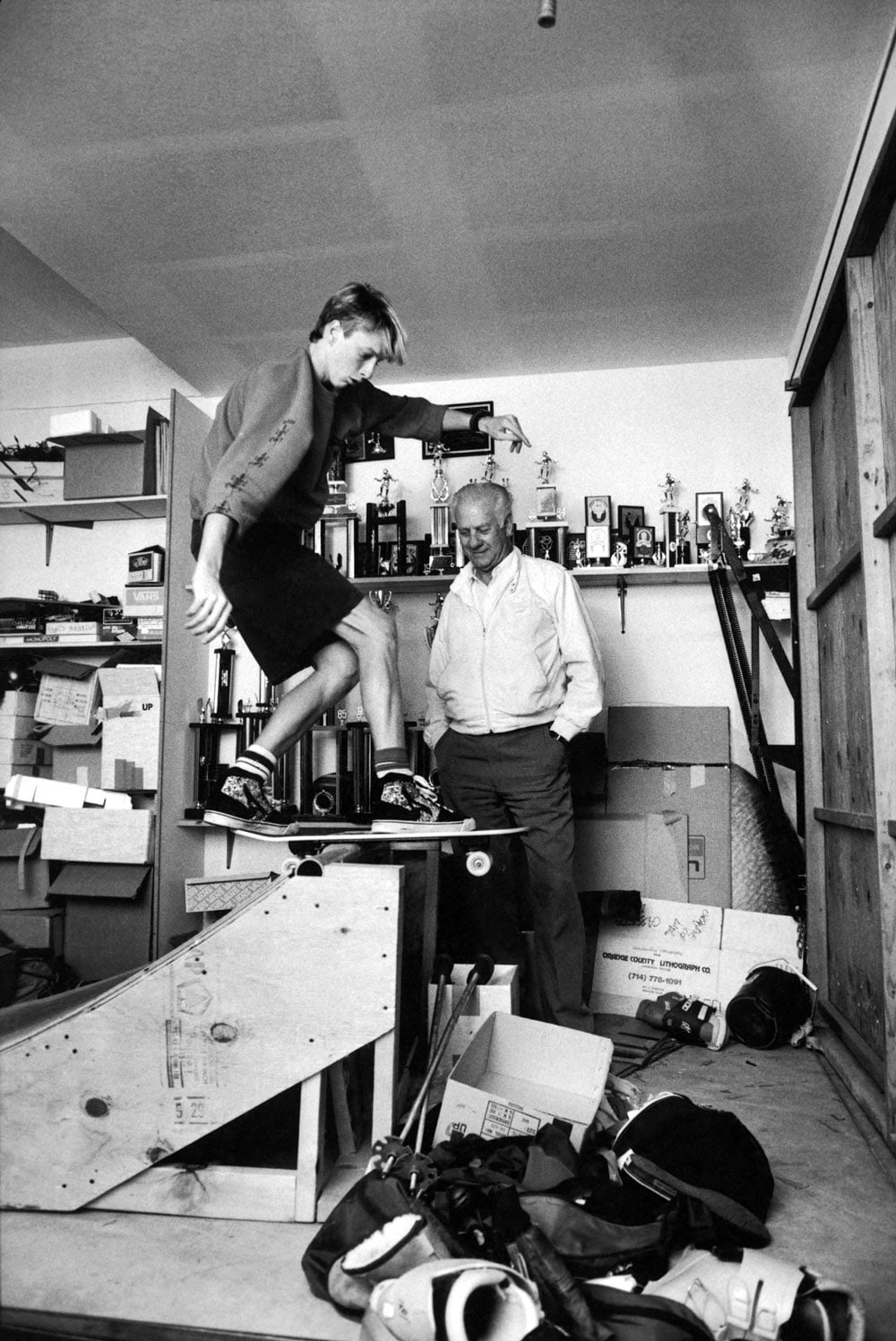 A teenage boy skates on a wooden ramp in a garage while an older man watches proudly, with a shelf full of trophies visible on the wall behind them.