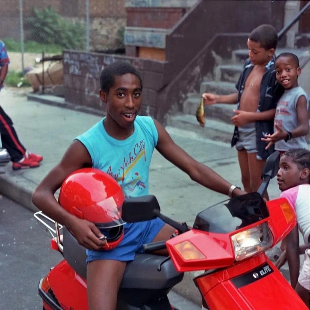A young Tupac on the back on a red scooter wearing a blue sleeveless shirt that says New York City 