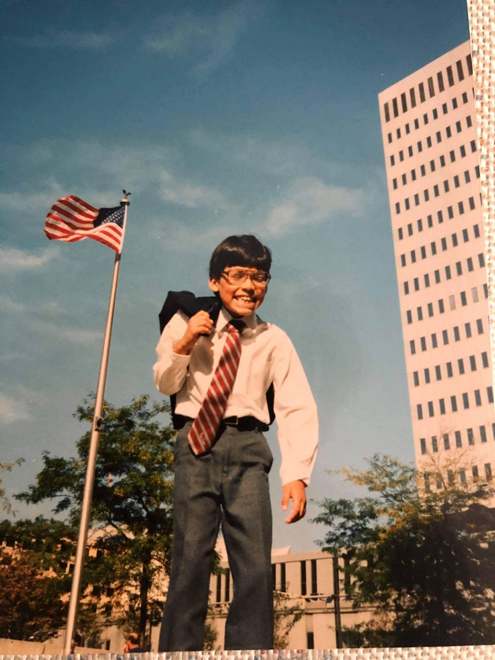 A beaming young boy in a white dress shirt, striped red and white tie, and slacks stands outside with an American flag flying behind him and a tall office building to his right.