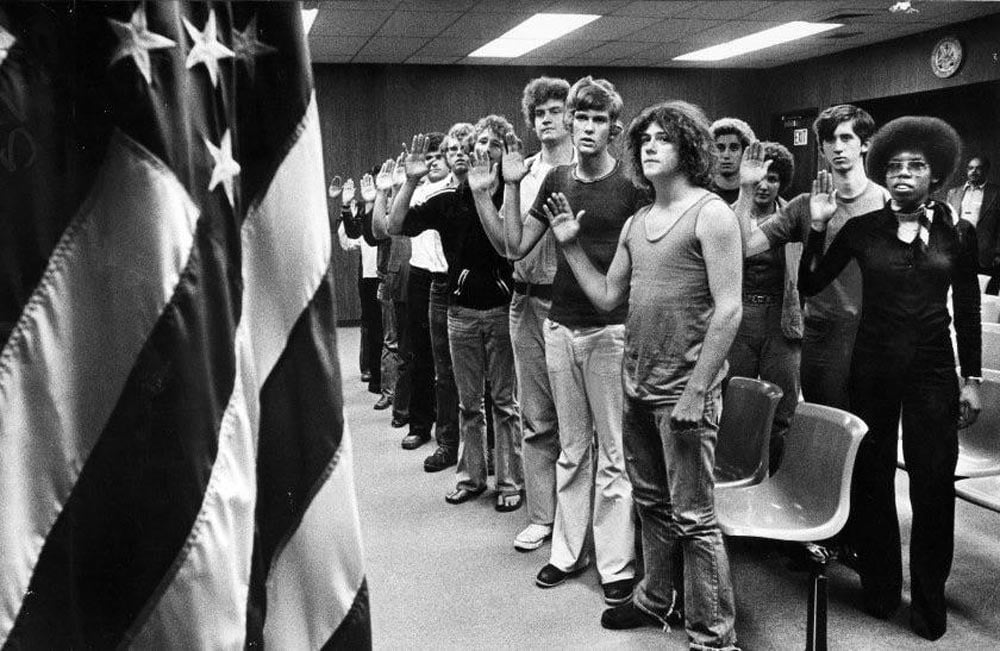 Black-and-white photo of a line of young men raising their right hands during an Army induction ceremony in Los Angeles, 1977. They wear casual civilian clothes including jeans, tank tops, and sandals. An American flag is visible in the foreground and a government seal hangs on the wood-paneled wall behind them.