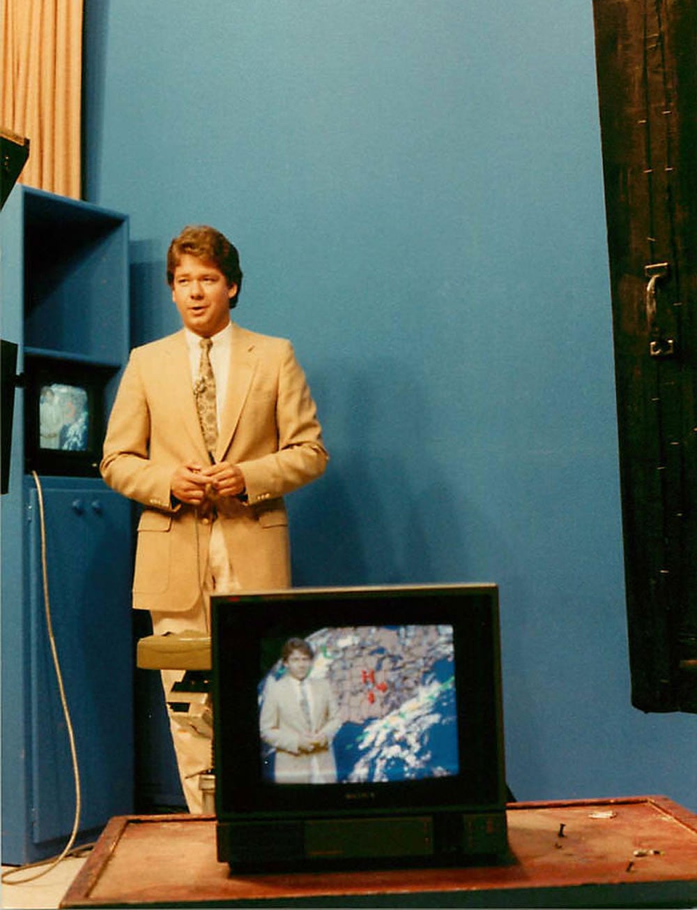 Man in a tan suit standing in front of a blue wall giving the tv weather report