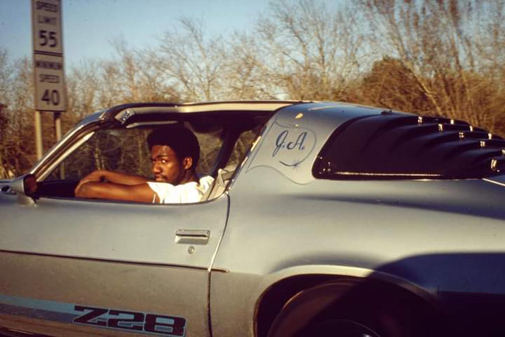 A looking back at the camera while behind the wheel of a silver Z28 Camaro