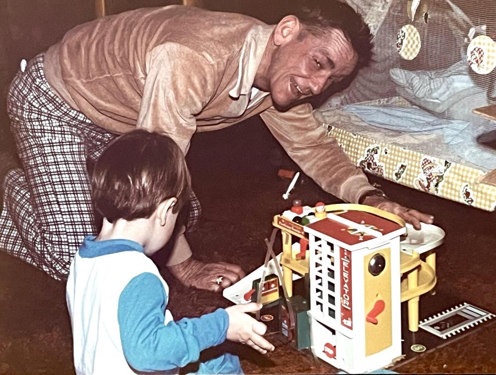 An older man on the floor playing with a kid and a fisher price parking garage. 