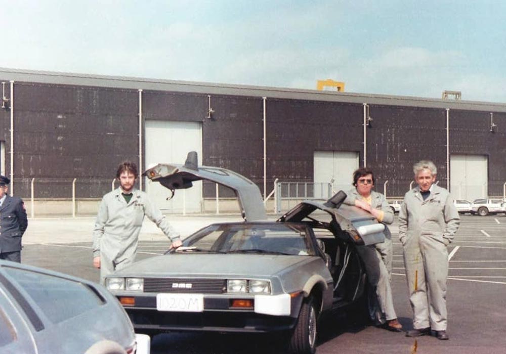 Three men in coveralls pose next to a DeLorean