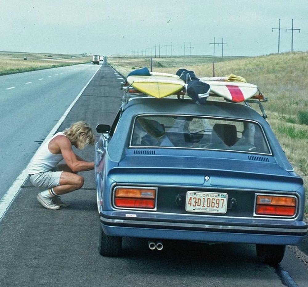 A woman working on his car on the side of the road with surf boards mounted to the roof