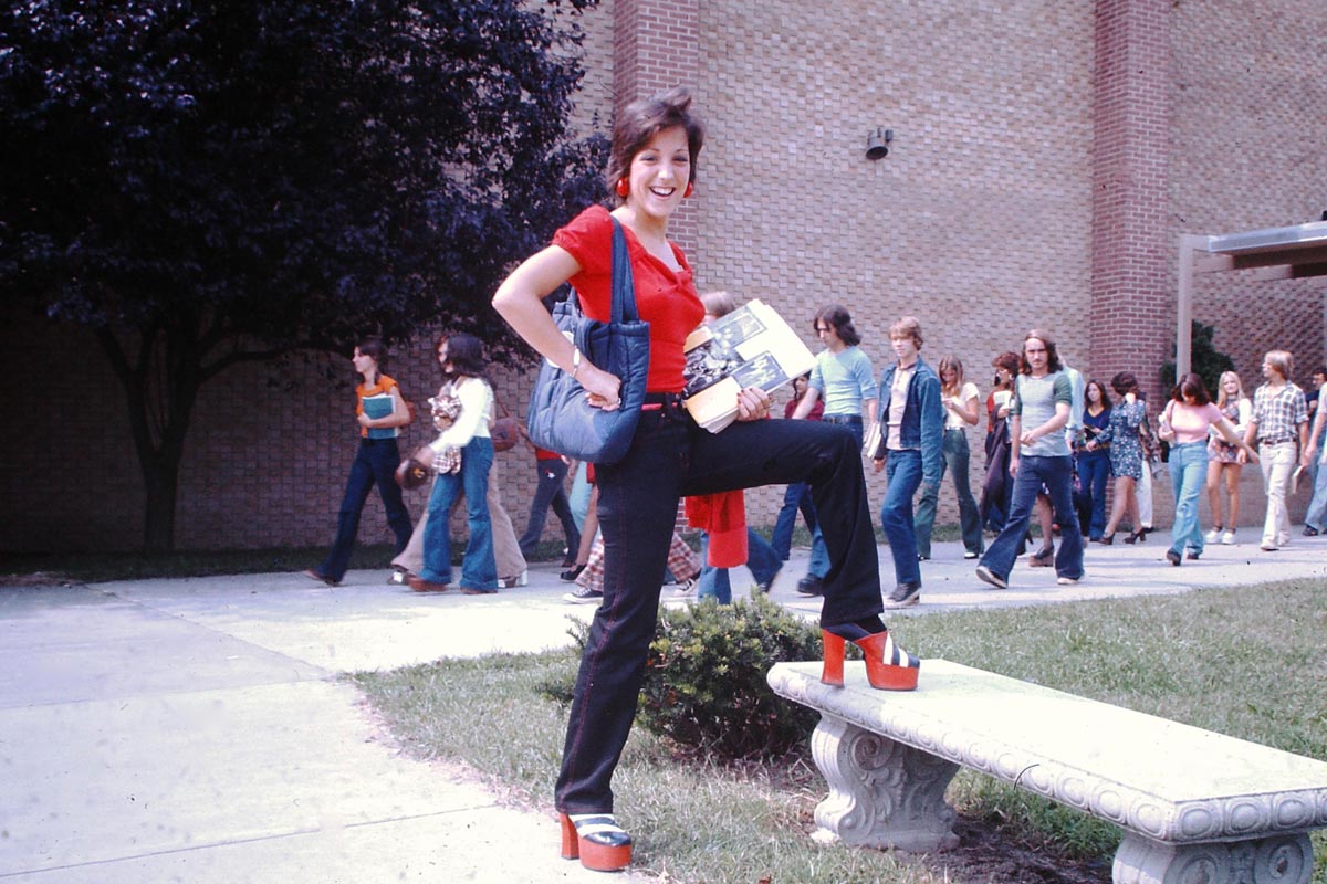 A teenage girl poses confidently outside her high school on the first day of school in 1975, one foot propped on a stone bench, wearing red platform shoes, black flared pants, and a red top, with students walking behind her.