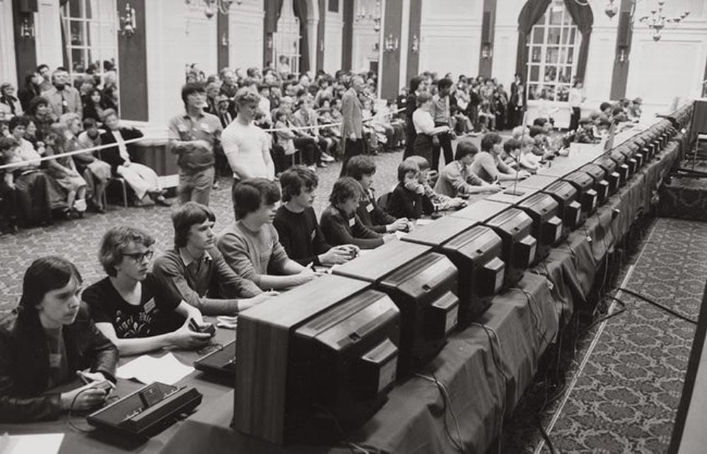Black and white photo of a row of kids playing space invaders in front of an audience
