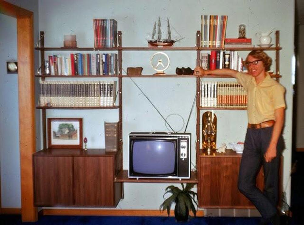 A young man standing in his livingroom next to a very small television
