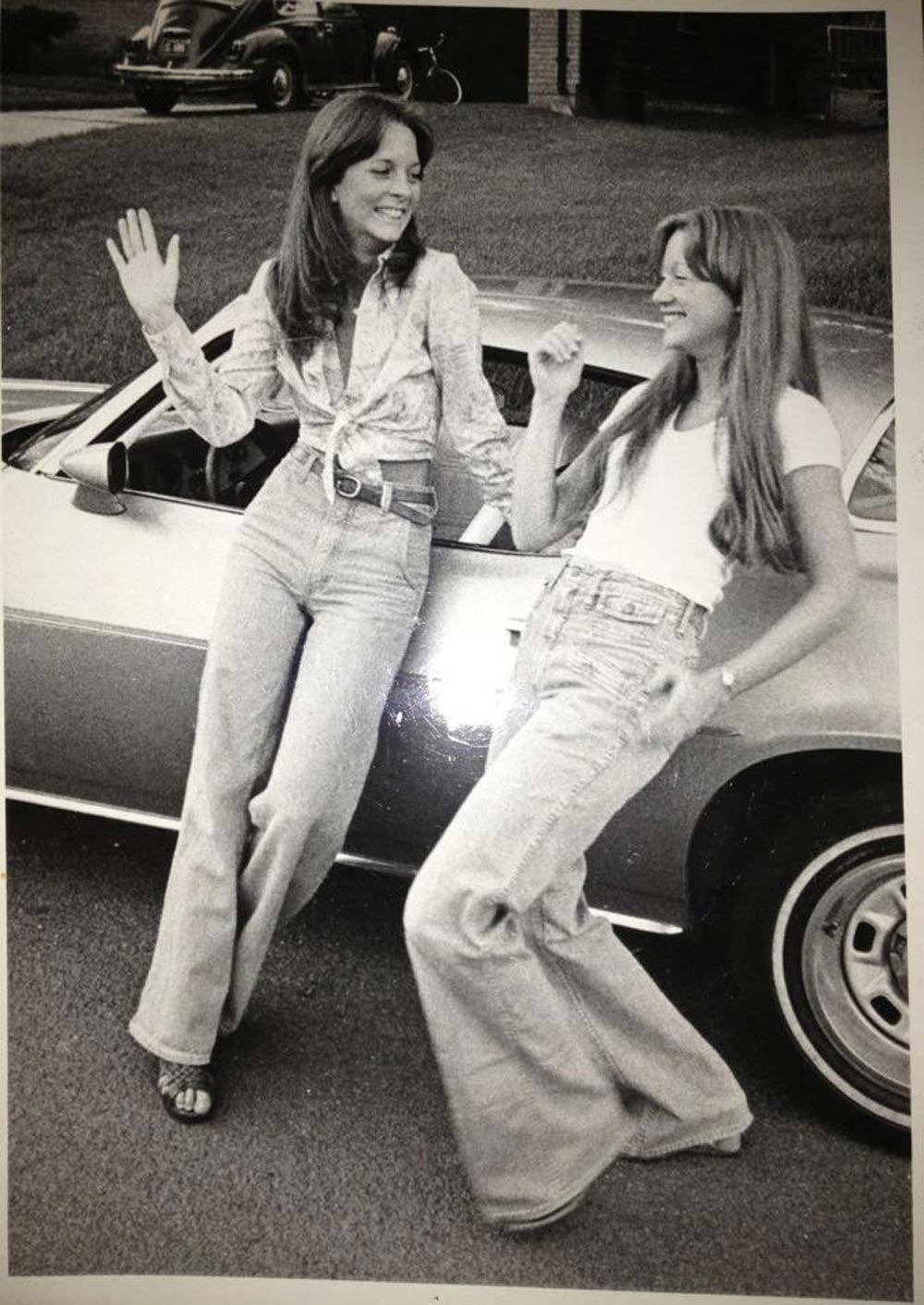 Black-and-white photo of two young women leaning against a car in a suburban driveway in 1977