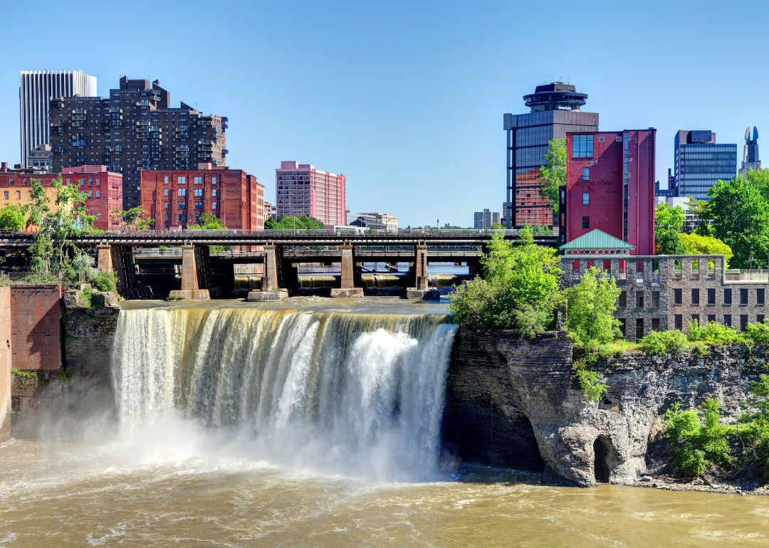 A waterfall under a bridge with the Rochester skyline in the background.