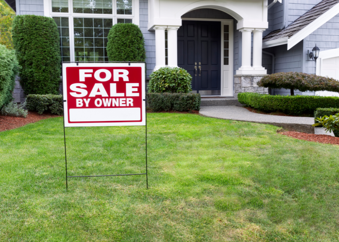 A for sale sign in the front lawn of a white suburban home.