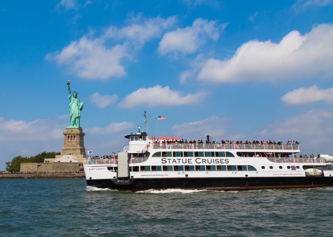 A crowded Statue Cruises tour boat in front of the Statue of Liberty.