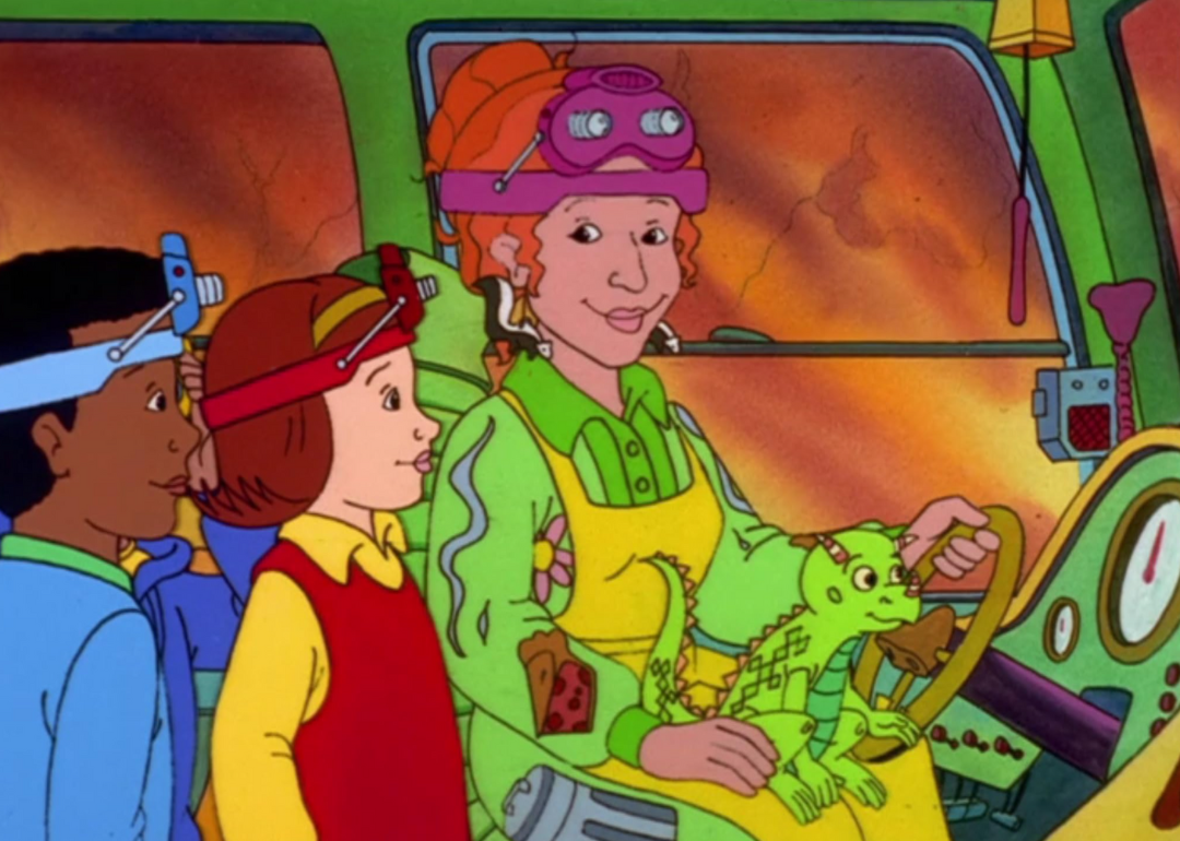Ms. Frizzle in the drivers seat of the bus with her lizard on her lap and two students standing next to her. 