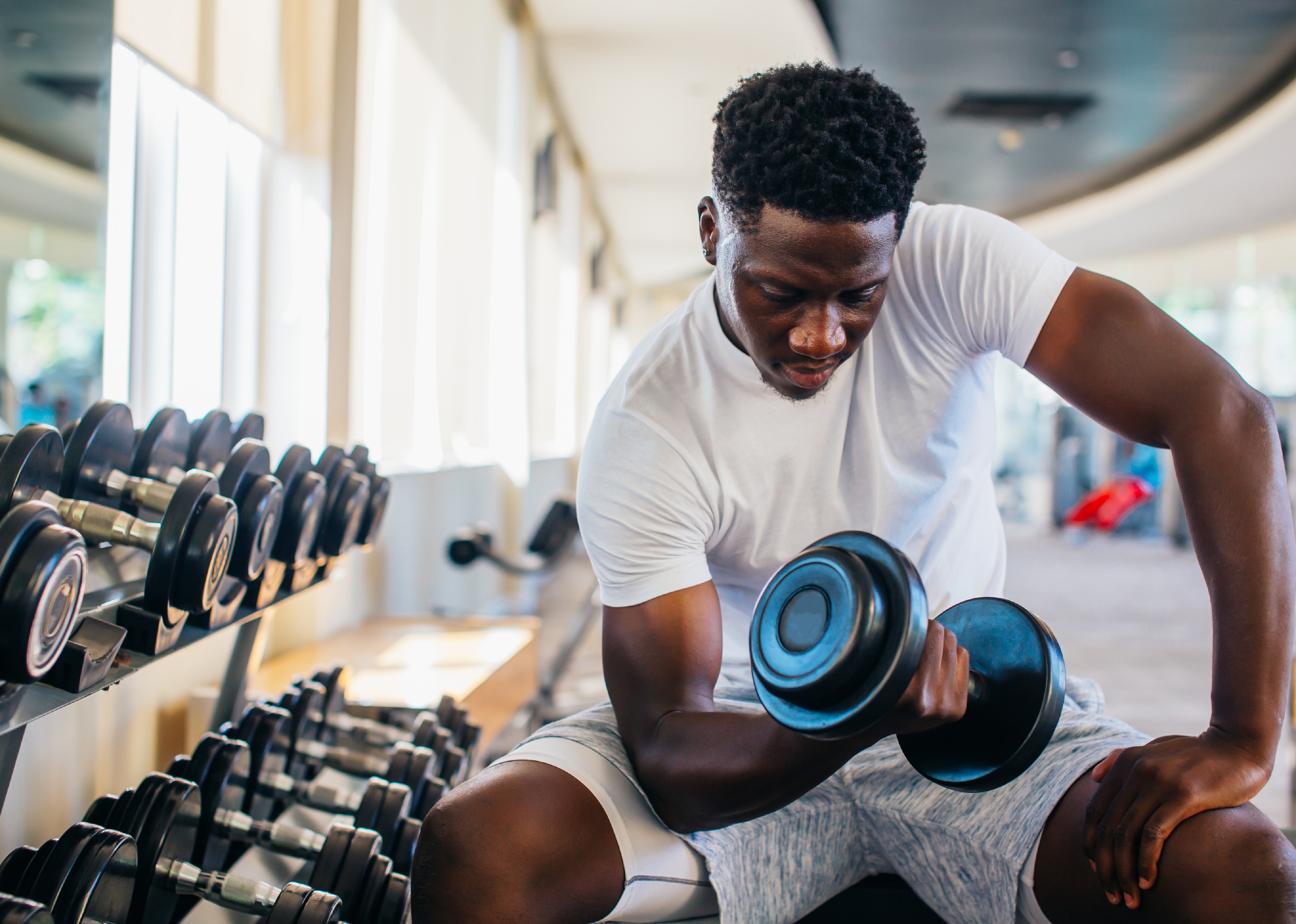 Young man sitting and lifting a dumbbell close to the rack at gym.