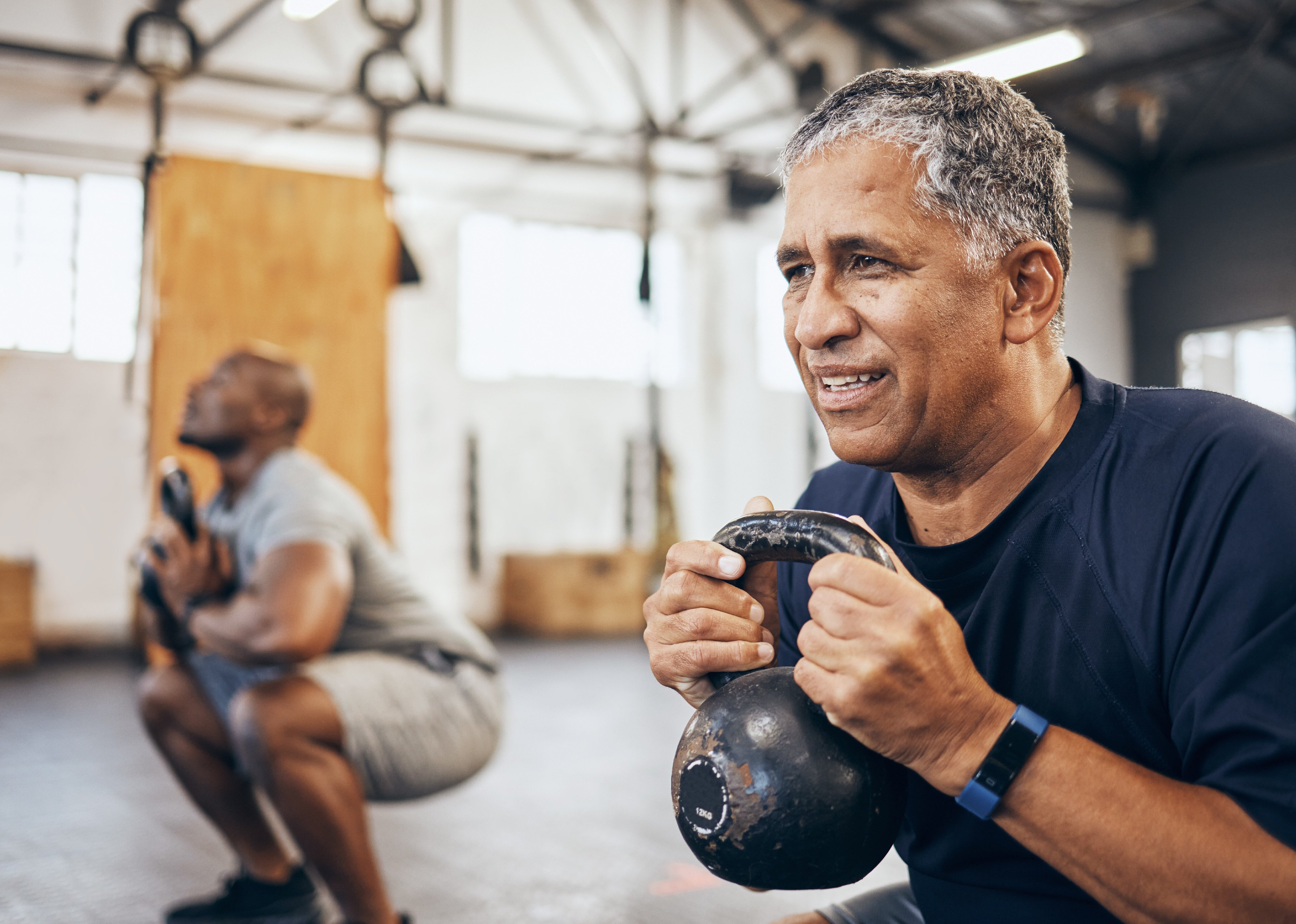 Two men squat with kettlebell equipment in a gym.