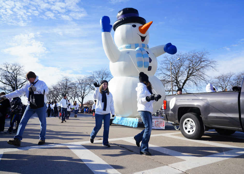 A holiday parade with people waving and a giant inflatable snowman being pulled by a pickup truck in Green Bay.