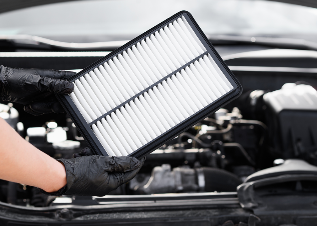 Hands holding a new air filter in front of a car engine.