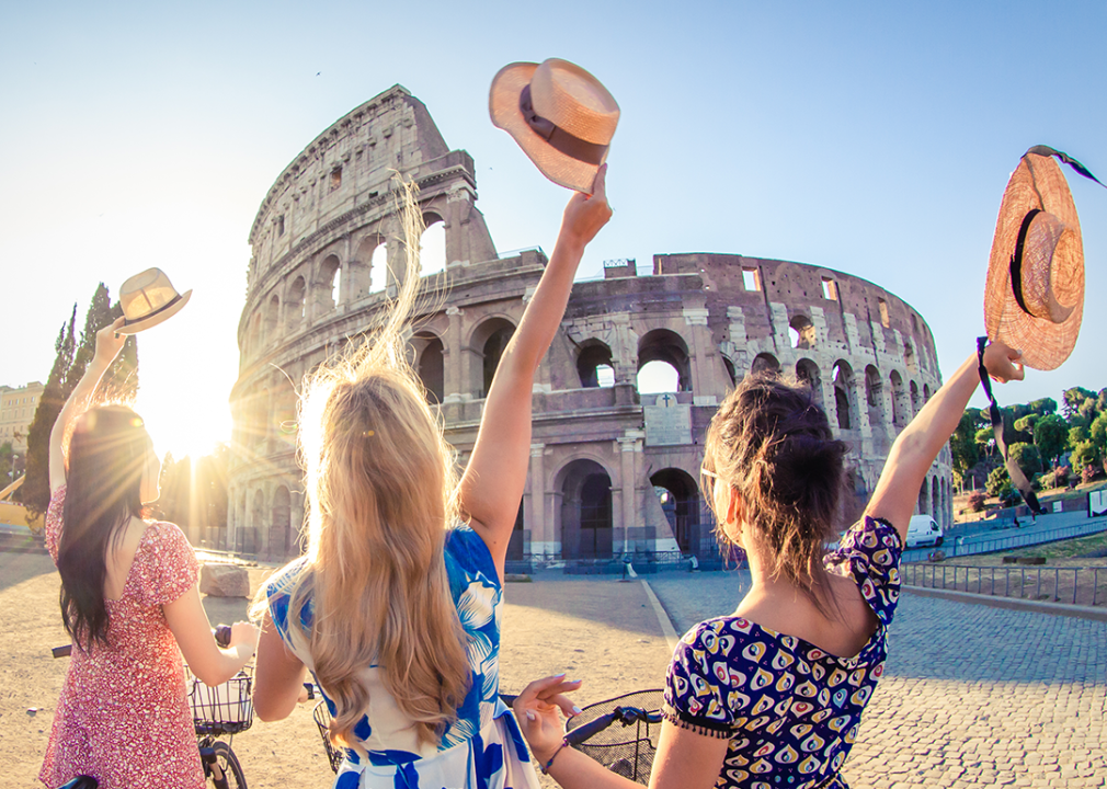 Three women wave hats at the Colosseum in morning sun.