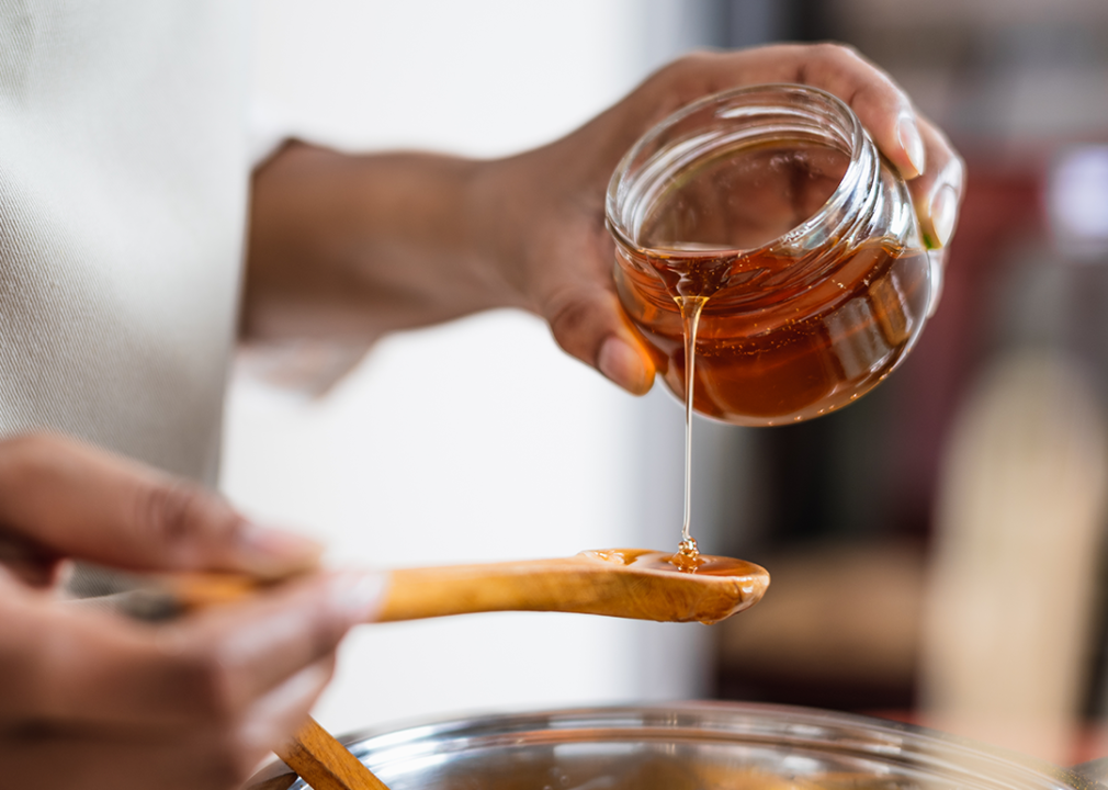 Hands pouring honey from jar to wooden spoon.